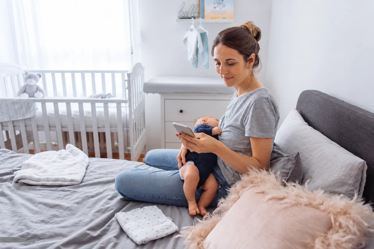Mother checking her phone while researching baby sleep consultant costs in the UK, sitting on bed with baby in lap and white crib visible in nursery background